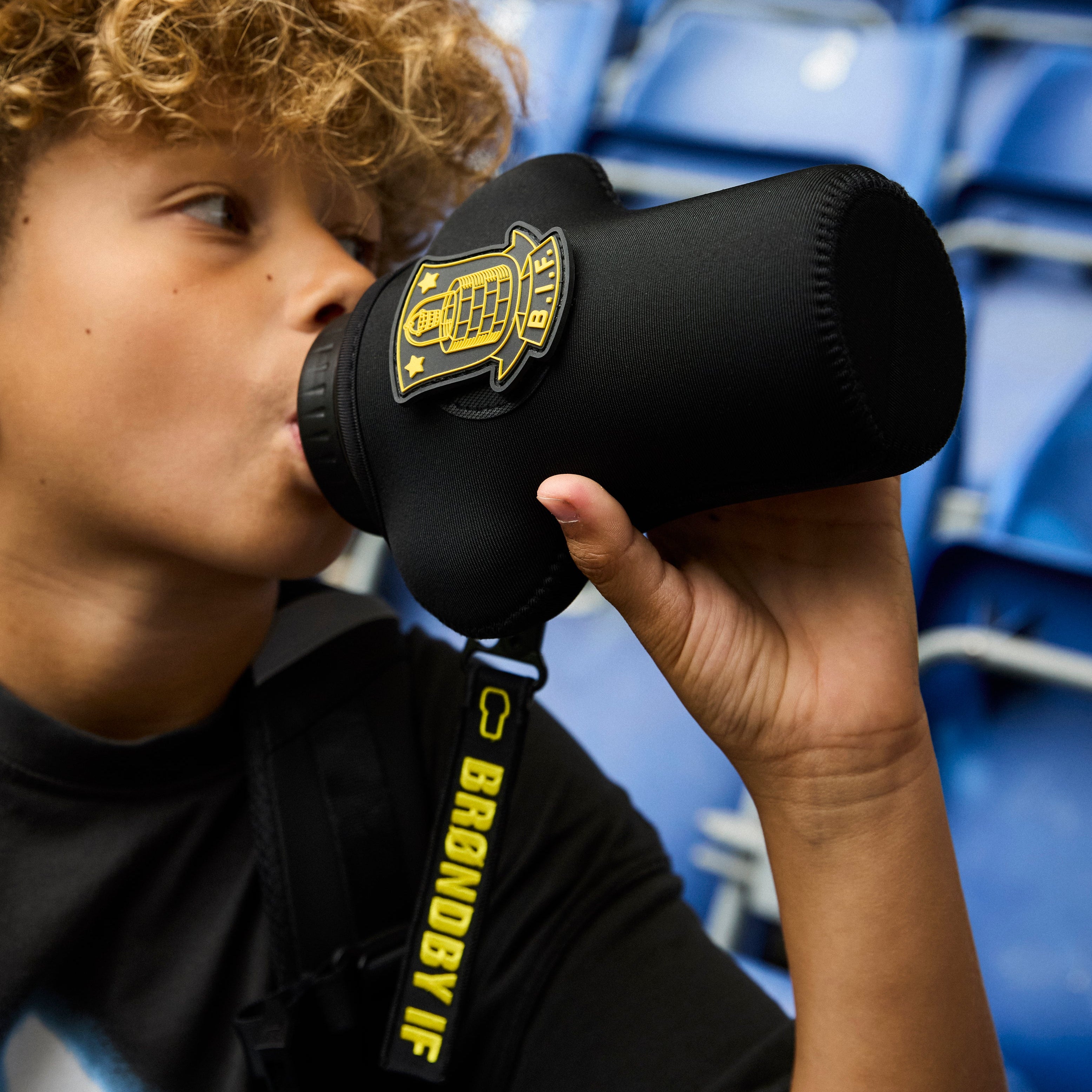 Person drinking from a black brøndby if water bottle in an stadium setting with blue seats.