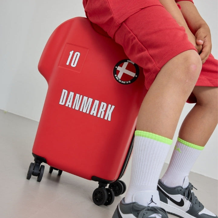 Person sitting on a red suitcase with 'Danmark' and number 10 printed on it, wearing red shorts and gray sneakers.