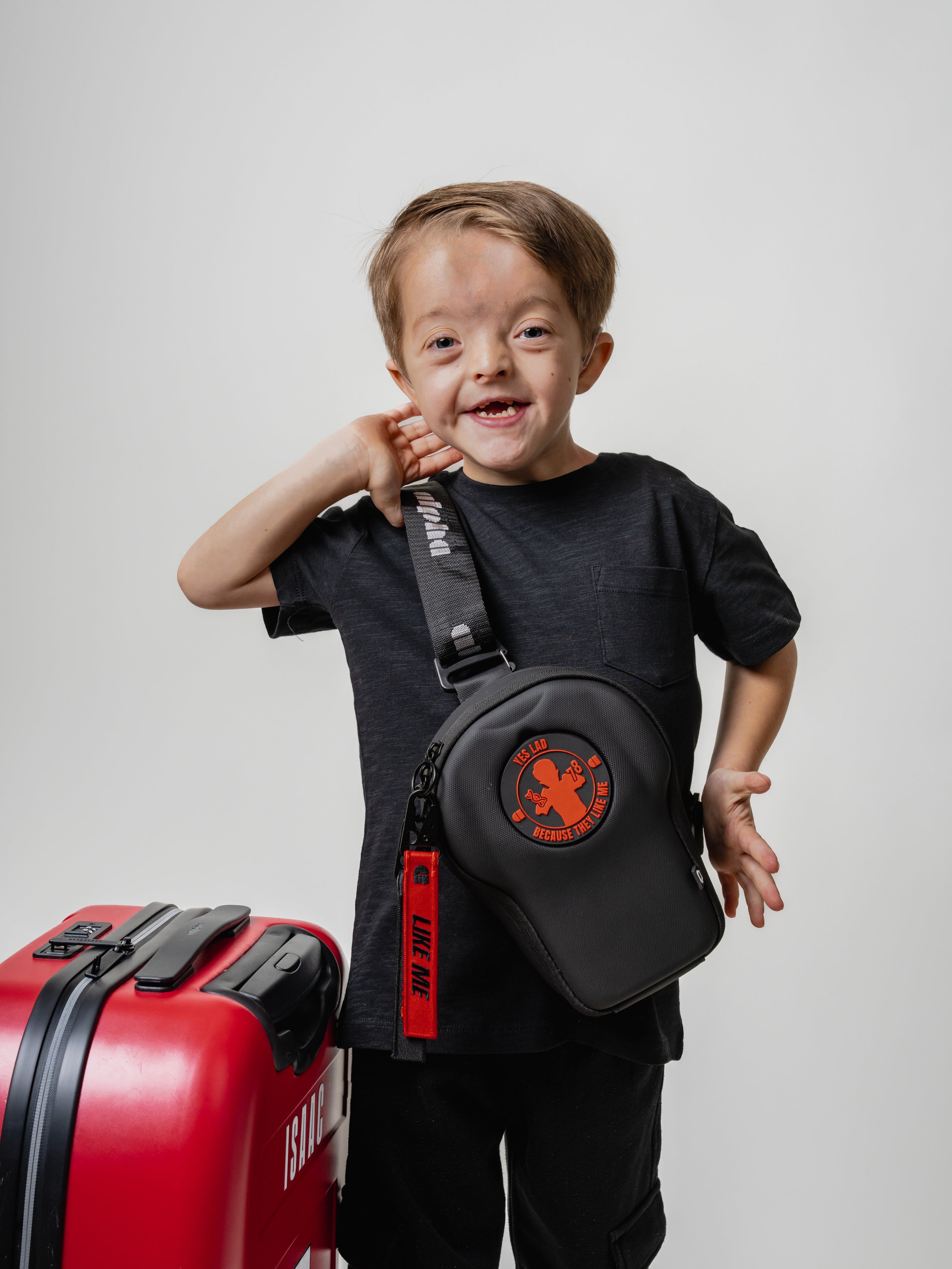 Child wearing a black sling bag with a red suitcase on a light gray background