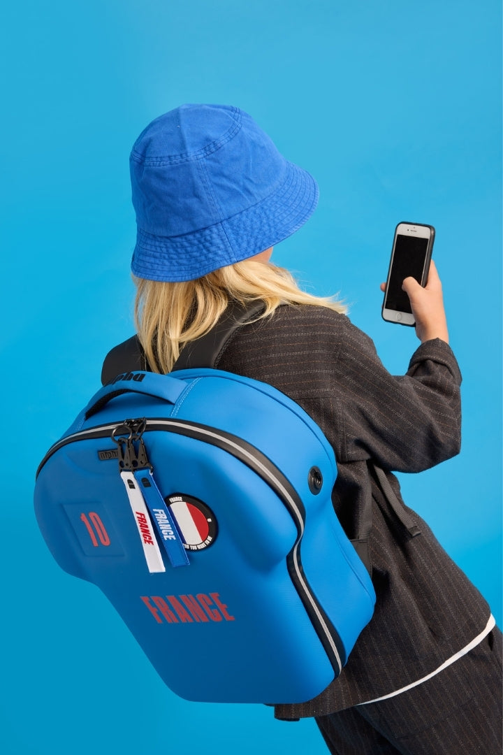 Person wearing a blue backpack with 'France' text and a matching blue hat, holding a phone on a solid blue background.