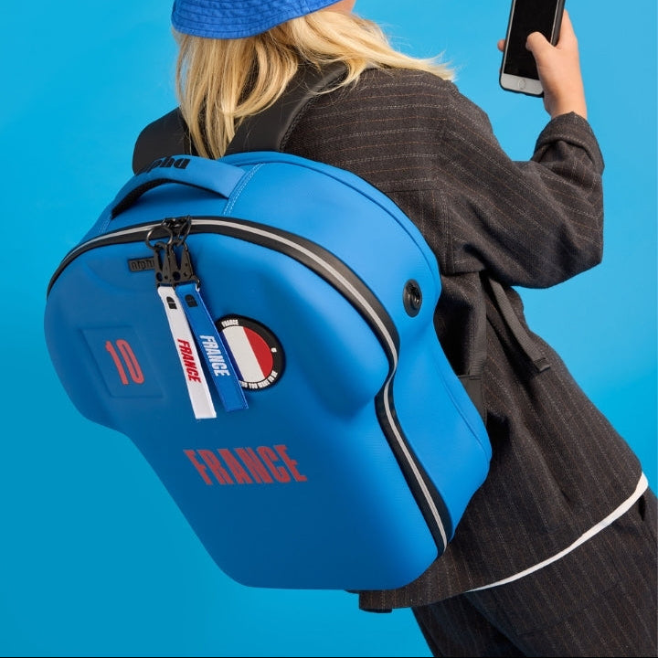 Person wearing a blue backpack with 'France' text and a matching blue hat, holding a phone on a solid blue background.
