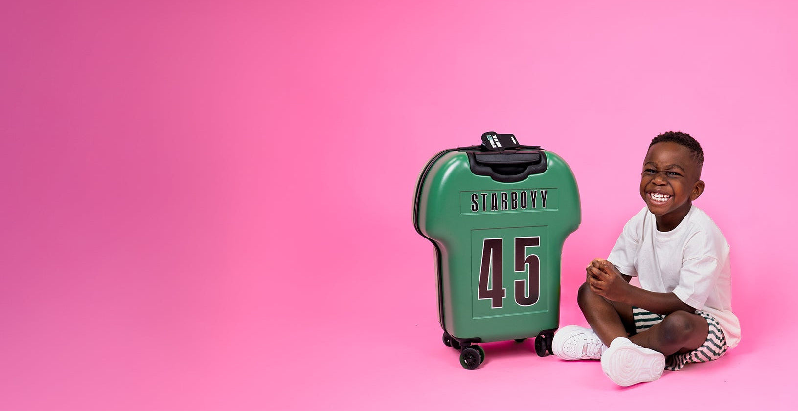 Kid sitting next to a green shirt-shaped cabin luggage with 'Starboyy' and number '45' on a pink background