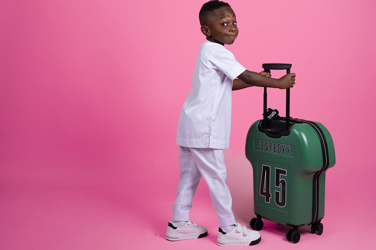 kid holding a green shirt-shaped cabin luggage with 'Starboyy' branding on a pink background by alpha