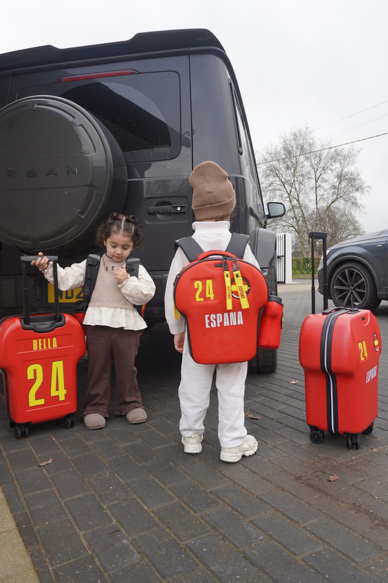 Two children with red suitcases labeled '24' and 'España' standing next to a black van.