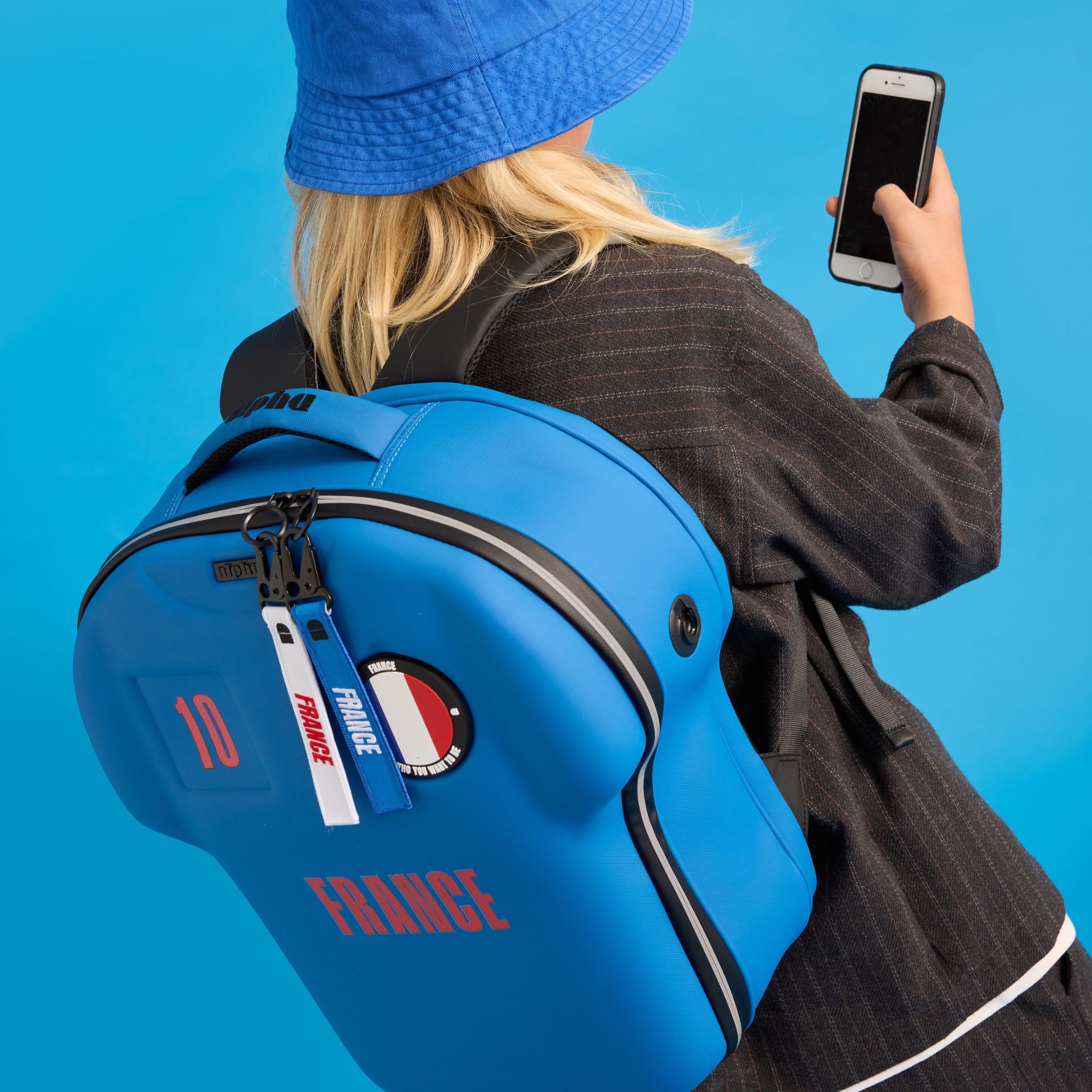 Person wearing a blue backpack with 'France' and flag design, holding a phone on a solid blue background