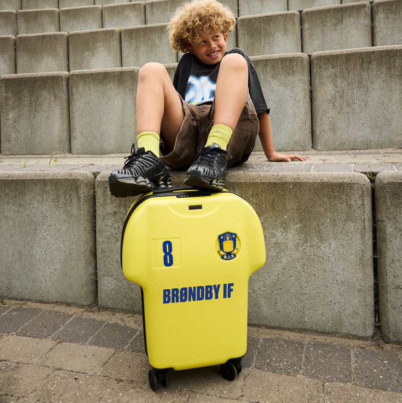 Child sitting on a yellow suitcase with 'BRONDBY IF' branding on concrete steps.