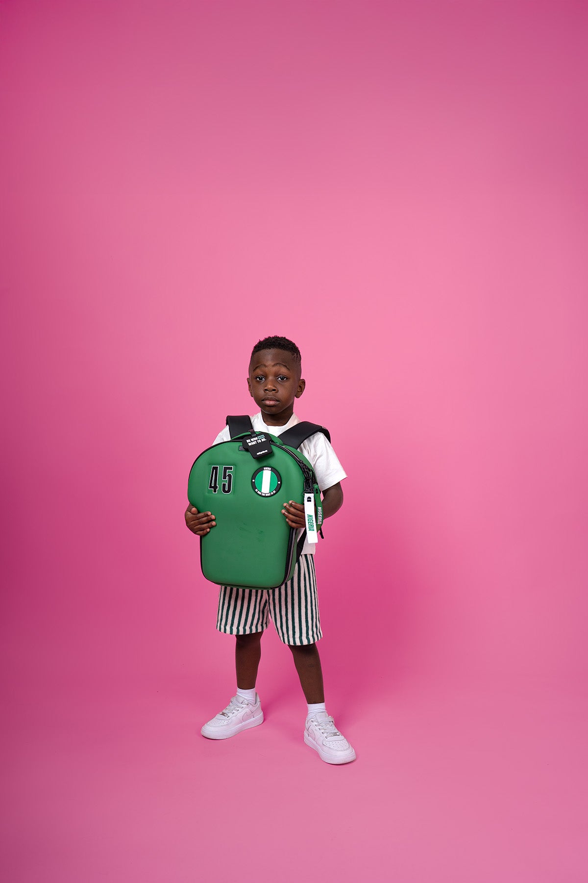 kid holding a green backpack against a pink background