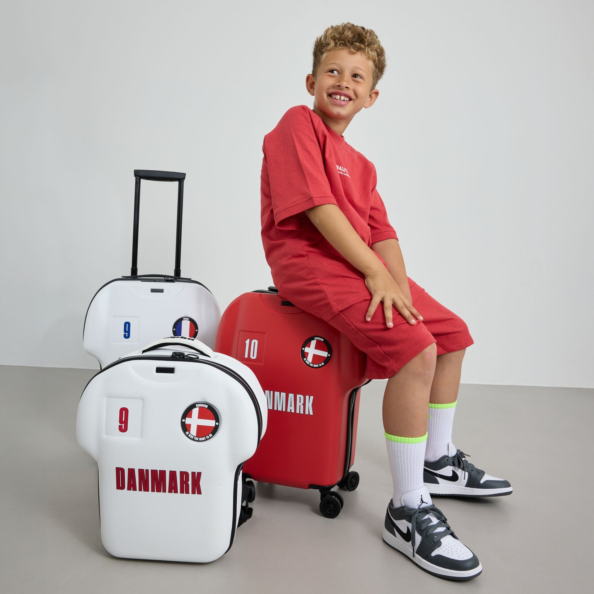 Child sitting on a red suitcase with 'Danmark' design, surrounded by white suitcases with Danish flags.