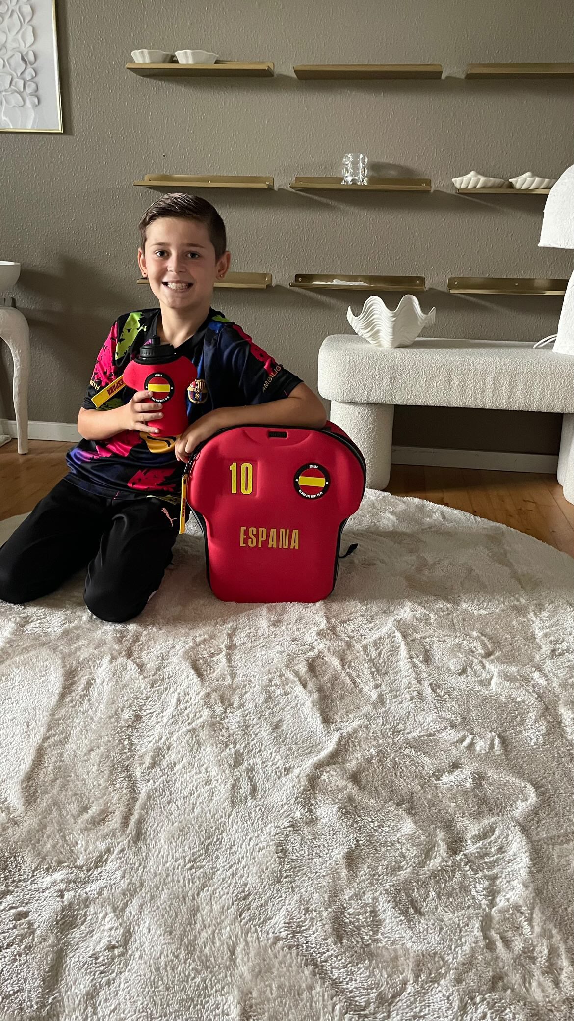 Child sitting on a rug with a red backpack labeled 'España' in a room.