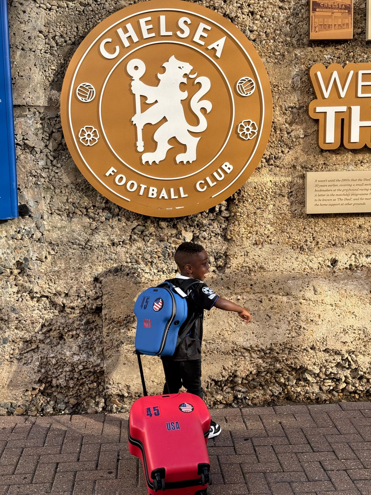 Child with a red cabin luggage and blue backpack standing in front of a Chelsea Football Club sign.